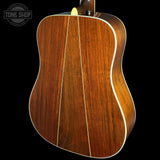 Close-up of the back of an acoustic guitar showing a rich reddish-brown wood grain finish with white binding, set against a solid black background. A Tone Shop watermark is visible in the top-left corner.