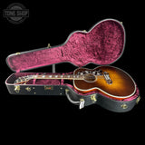 A sunburst acoustic guitar resting inside an open black hard case with magenta-pink velvet lining, shown against a solid black background. A Tone Shop watermark is visible in the top-left corner.