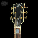 Close-up of a Gibson acoustic guitar headstock featuring gold tuning pegs, a black faceplate with white script logos, and a dark wood fretboard against a black background.