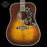 A close-up of an acoustic guitar body with a natural sunburst finish, featuring a dark brown pickguard decorated with white and yellow bird and floral inlays, set against a solid black background.