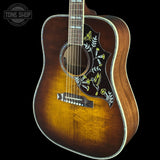 A close-up of an acoustic guitar body showing a natural wood sunburst finish, a black pickguard with yellow and white floral inlay, and a white binding edge, set against a solid black background with a Tone Shop watermark in the top left.