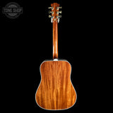 The back of a wooden acoustic guitar, showing a rich cherry wood grain finish, photographed against a solid black background with a Tone Shop watermark in the top-left corner.