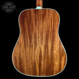Close-up of the back of an acoustic guitar showing a rich, warm brown wood grain finish with a white edge trim, set against a solid black background.