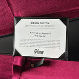 A Gibson Custom Certificate of Authenticity card inside a black frame, placed on a deep magenta velvet-lined Gibson instrument box.