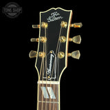 Close-up of the headstock of a Gibson Anniversary electric guitar against a solid black background. The headstock is black with gold hardware and features the Gibson logo and 'Anniversary' inscript on the pickguard.