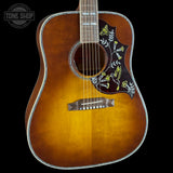 A close-up of an acoustic guitar body with a natural sunburst finish, featuring a dark brown pickguard decorated with yellow and white floral inlay, set against a solid black background.