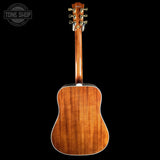 The back of a Gibson acoustic guitar with a rich, warm brown wood finish, shown against a solid black background with a Tone Shop watermark in the top left corner.