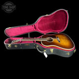 An acoustic guitar with a sunburst finish displayed inside an open black hard case with bright pink velvet lining, set against a solid black background. A Tone Shop watermark is visible in the top-left corner.
