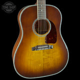 A close-up of an acoustic guitar body showing a classic sunburst finish that transitions from a bright golden yellow center to dark brown and black edges. The guitar features a dark brown pickguard, a black bridge, and a silver-toned metal truss rod cover.