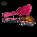 An acoustic guitar with a sunburst finish displayed inside an open black hard case with a bright pink velvet interior, set against a solid black background. A Tone Shop watermark is visible in the top-left corner.
