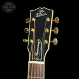 Close-up of the headstock of a Gibson guitar, featuring gold tuning pegs, a black headstock with white script reading 'The Gibson' and 'Anniversary', and a dark fretboard against a black background.