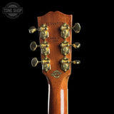 Close-up of the headstock of a wooden acoustic guitar against a black background, showing six gold-toned tuning pegs and printed details including '21346929' and 'MADE IN U.S.