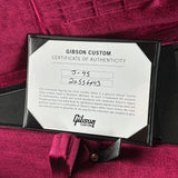 A close-up of a Gibson Custom Certificate of Authenticity card in a black frame, placed on a deep magenta velvet fabric background with part of a black guitar headstock visible.