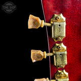 Close-up of a guitar headstock showing two gold-toned tuning pegs and matching gold-toned tuning machines against a black background, with part of a red wooden body visible. A Tone Shop watermark is in the upper left corner.