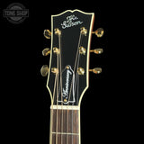 Close-up of the headstock of a Gibson Anniversary acoustic guitar against a black background, featuring gold tuning pegs, white script logos, and a dark wood fretboard.
