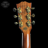 Close-up of the headstock of a Gibson acoustic guitar in a warm brown finish, featuring six gold-toned tuning pegs and a black background.