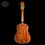 The back of a wooden acoustic guitar, showing the rich grain of the mahogany-style backplate and headstock, photographed against a solid black background with a Tone Shop watermark in the top left corner.