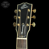 Close-up of the headstock of a Gibson acoustic guitar, featuring gold tuning pegs, a black headstock with white script, and a white fretboard against a black background.