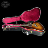 A Gibson acoustic guitar with a sunburst finish displayed inside an open black hard case with a magenta-pink velvet interior, set against a solid black background.