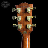 Close-up of the headstock of an electric guitar, showing six gold-toned tuning pegs, a glossy brown wood finish, and a circular Gibson logo near the bottom, set against a solid black background.