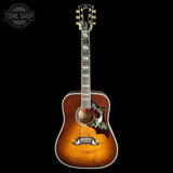 A vintage-style acoustic guitar with a sunburst finish, featuring a floral inlay on the lower bout, set against a solid black background.