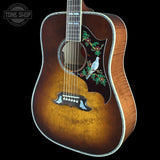 A close-up of an acoustic guitar body against a black background, featuring a natural sunburst finish that transitions from dark brown to golden amber. The pickguard is black and decorated with a detailed illustration of a white bird perched on a green and red floral branch.