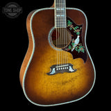 A close-up of a vintage acoustic guitar body with a natural sunburst finish, featuring a decorative pickguard with a white bird and green floral inlay, set against a solid black background.