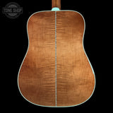 The back of a wooden acoustic guitar, showing a rich flamed maple top finish in warm brown tones with a light blue-green binding edge, set against a solid black background.