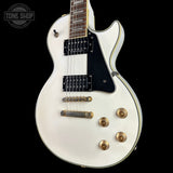 A white electric guitar with a black pickguard and gold hardware, shown against a solid black background. A Tone Shop watermark is visible in the top-left corner.