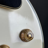 Close-up of a white electric guitar body with a gold-toned volume control knob and part of a black pickguard.