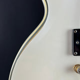 Close-up of a white electric guitar body showing the edge of the body, a gold-toned binding strip, and part of a black hardware component against a dark background.