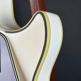 Close-up of an electric guitar body showing a cream-colored finish with black and gold binding, set against a dark gray background.