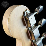 Close-up of the headstock rear corner of a guitar with a light natural wood backplate and silver tuning pegs against a black background. A Tone Shop watermark is visible in the top-left corner.