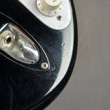 Close-up of a glossy black electric guitar body with a chrome headlight cover, white control plate, and white volume knob against a neutral gray background.