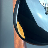 Close-up of a glossy black electric guitar body showing a small oval wood inlay and part of a white pickguard, with a blurred gray and wood background.