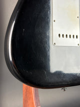 Close-up of the glossy black body of an electric guitar, showing a worn silver plate, scratches, and part of the wooden neck.