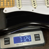 A close-up of a digital scale displaying the weight of a black electric guitar, with the scale showing 7.124 oz and part of the guitar's white pickguard and neck visible.