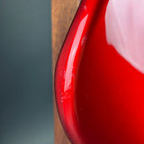 Close-up of a glossy red teardrop-shaped object, likely a decorative vase or bowl, showing a curved edge with a reflective finish against a dark gray and wood background.