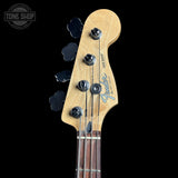 Close-up of a Fender Jazz Bass headstock with a natural maple wood finish, four black tuning pegs, and the blue Fender logo, shown against a solid black background.