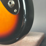 Close-up of the side of a sunburst electric guitar body showing a glossy black edge, silver hardware, and a faded amber-to-brown finish against a soft gray background.