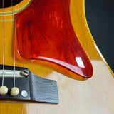 Close-up of a guitar body showing a glossy reddish-brown pickguard over a natural wood finish, with part of the bridge and strings visible.