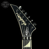 Close-up of a Jackson electric guitar headstock in black with gold lettering, shown against a solid black background. The Jackson brand name is written in gold script on the headstock, with tuning pegs and strings visible.