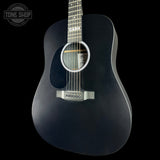 A black acoustic guitar with a dark wood neck and silver hardware, shown against a solid black background. A circular 'TONE SHOP' watermark is visible in the top-left corner.