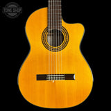 A classical-cutaway acoustic guitar with a natural golden-yellow spruce top, dark fretboard, and black pickguard, shown against a solid black background with a Tone Shop watermark in the top left corner.