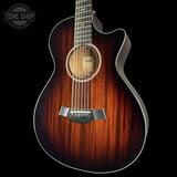 A Taylor acoustic guitar with a dark brown sunburst finish, shown from a front-three-quarter angle against a solid black background. The brand logo 'TONE SHOP' is visible as an overlay in the top-left corner.