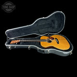 An acoustic guitar with a natural wood finish resting inside an open black hard case on a solid black background. A Tone Shop watermark is visible in the top-left corner.