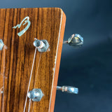 Close-up of a wooden guitar headstock with visible wood grain, a stylized logo plaque, and metal tuning pegs against a dark blue background.