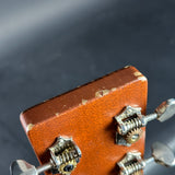 Close-up of the headstock of a guitar, showing a textured brown wood finish with visible wear and scratches, and silver metal tuning pegs against a dark gray background.