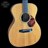 A close-up studio shot of an acoustic guitar against a solid black background, featuring a natural wood finish, a dark fretboard, and a decorative pickguard.