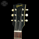 Close-up of a Gibson Les Paul TV Model acoustic guitar headstock, featuring a black finish with gold lettering and cream-colored tuning pegs, set against a solid black background.